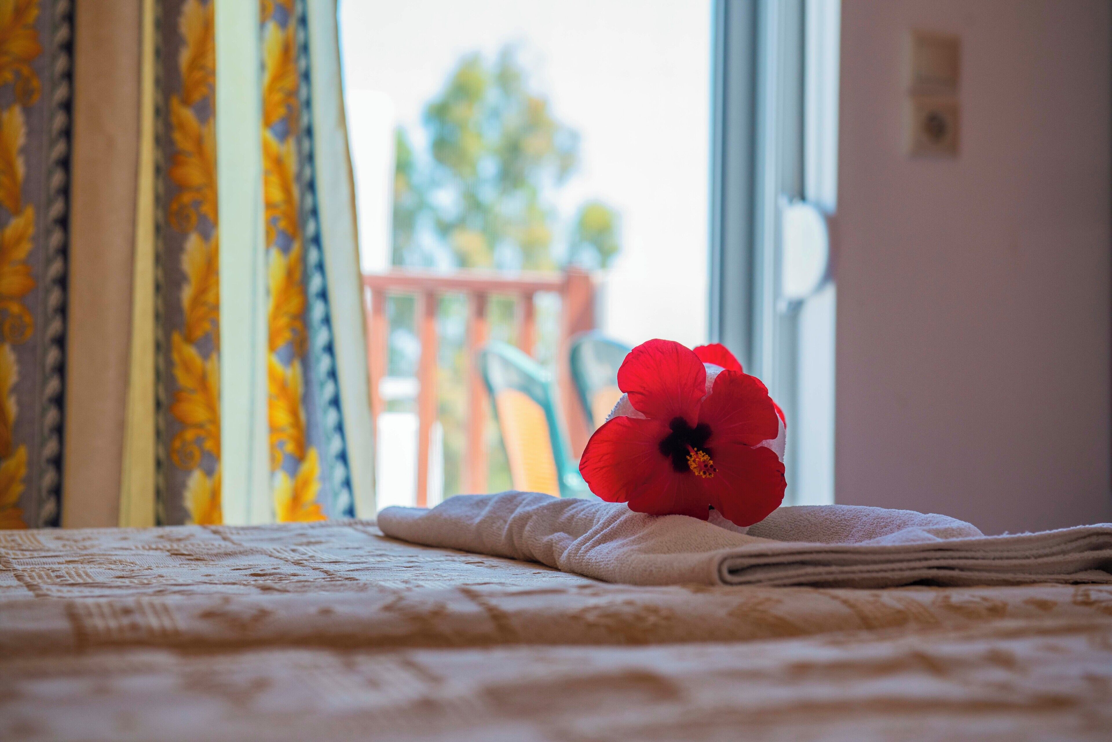 Un lit avec une serviette roulée et une fleur rouge, des rideaux à motifs floraux et une vue sur un balcon avec une balustrade.