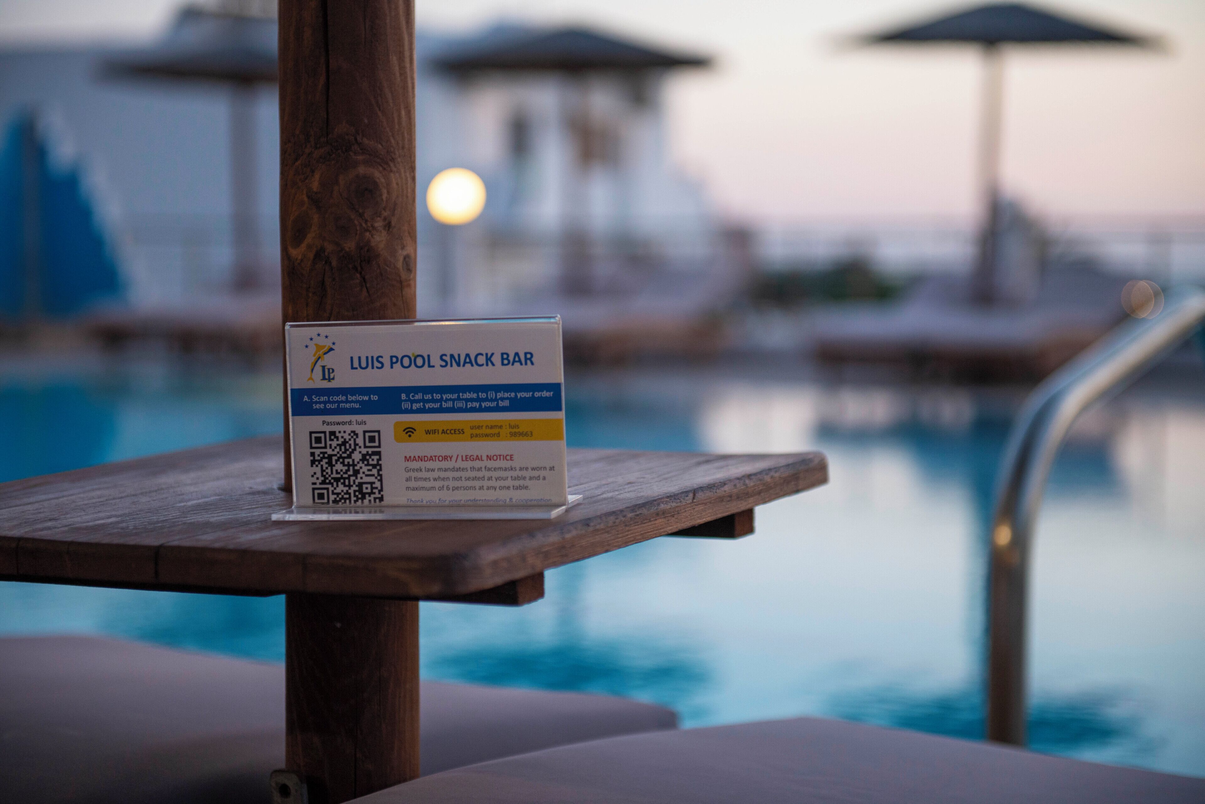 A QR code sign for Luis Pool Snack Bar with a pool in the background.
