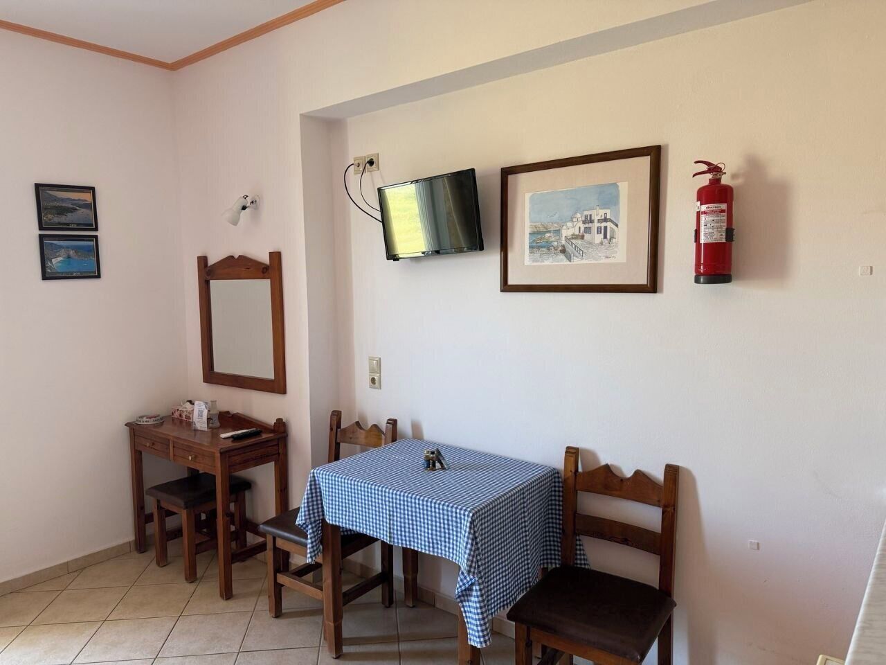 A dining area with a table covered in a checkered tablecloth, wooden chairs, a wall-mounted television, a framed picture, and a fire extinguisher.
