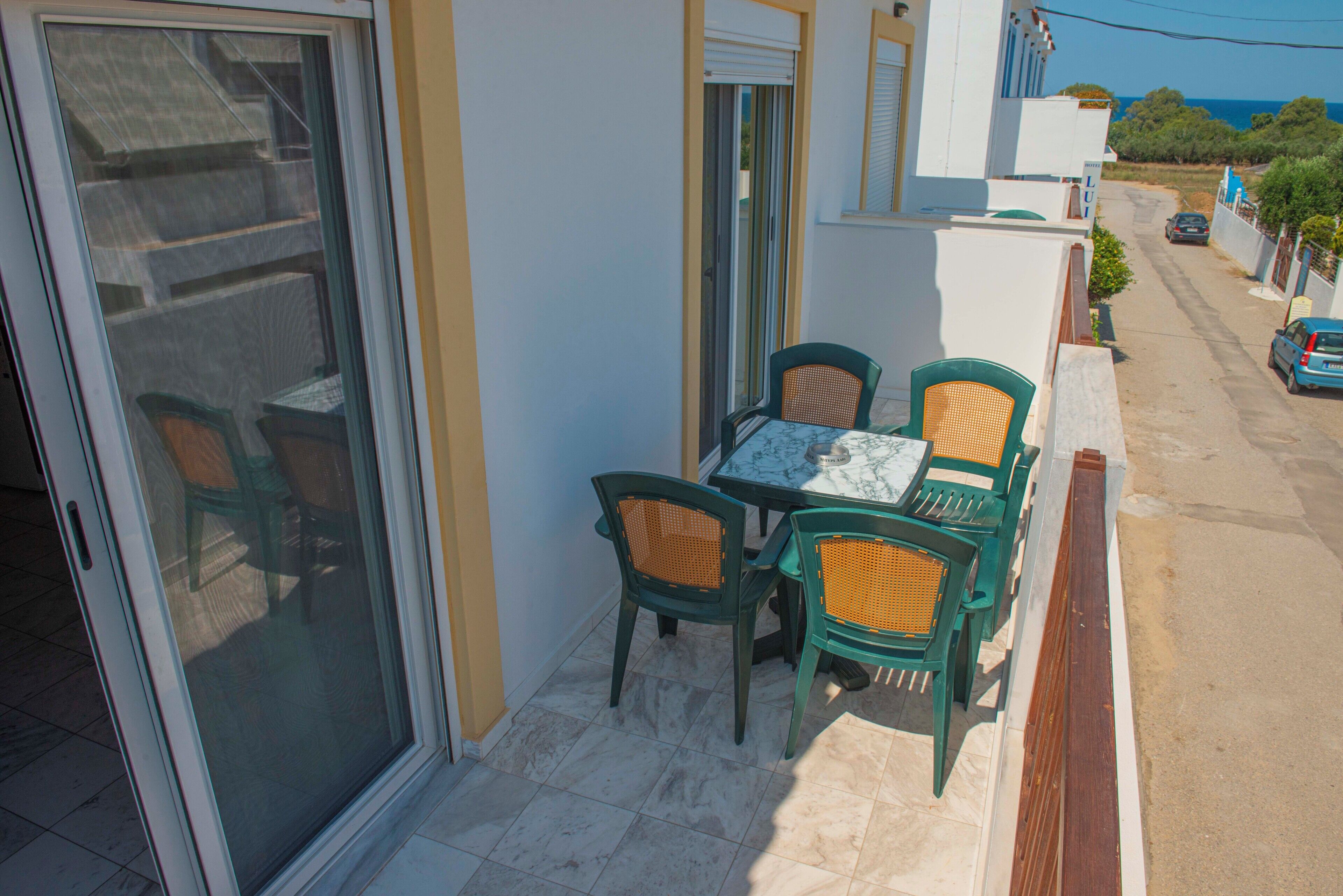 A balcony with a table and chairs, a sliding glass door, and a view of a street and ocean.