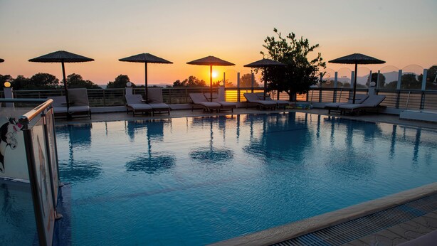 A swimming pool with lounge chairs and umbrellas, surrounded by a low fence, with a sunset in the background. - Luis Pool (Kos)