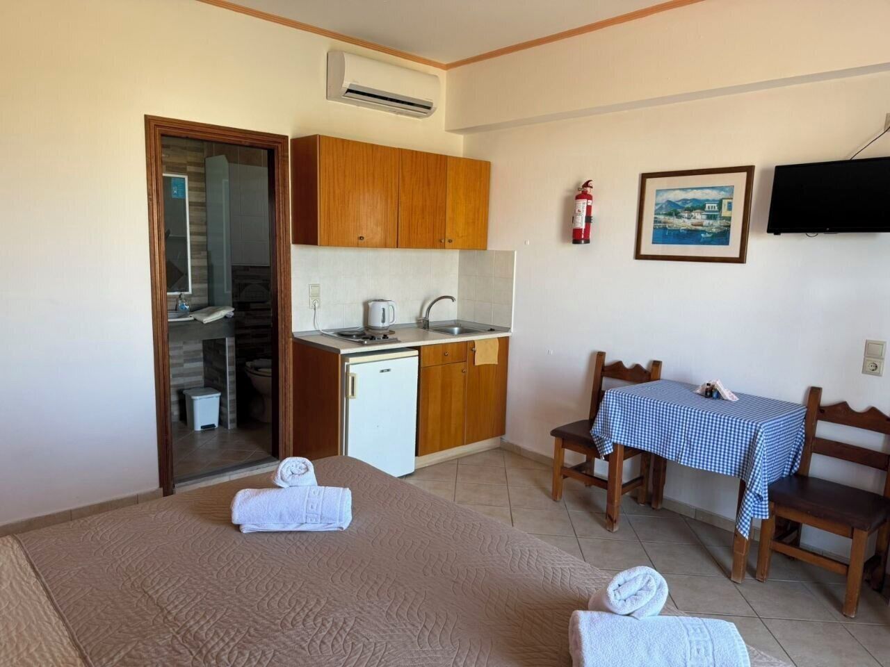 A room with a bed, a small kitchenette, a table with a checkered tablecloth, and a TV on the wall.
