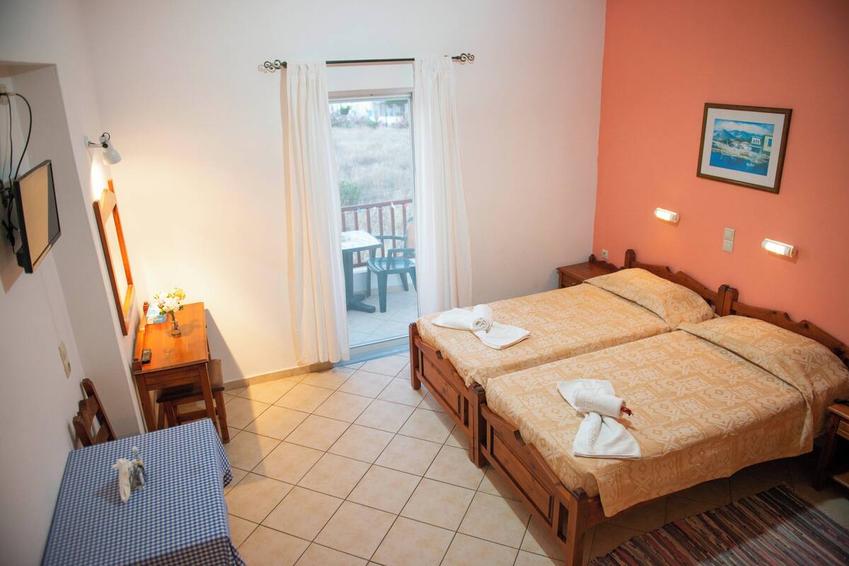 A hotel room with two beds, a dining table, a chair, a lamp, and a view of the outside.