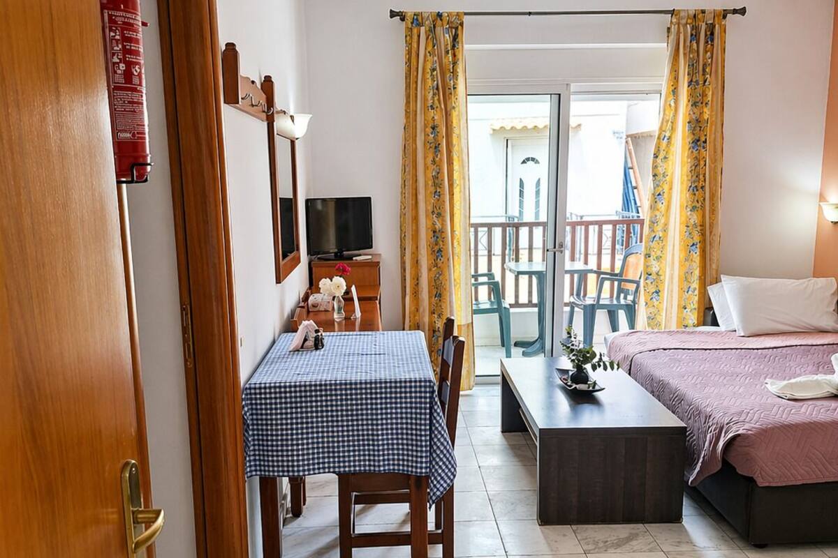 A room with a checkered table, a bed, a television, and a balcony with yellow curtains.