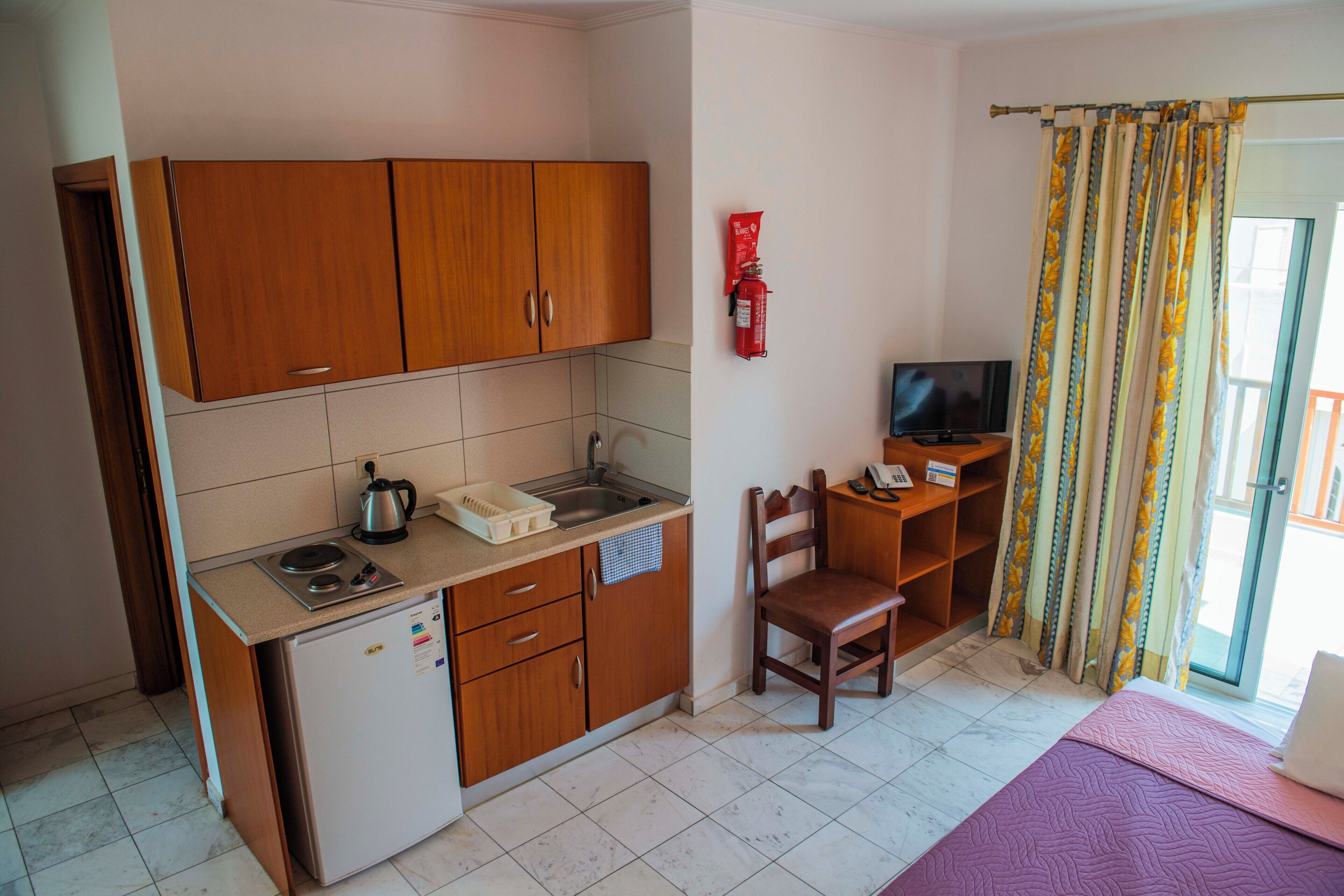 A compact kitchen with wooden cabinets, a small refrigerator, a sink, and a kettle. There's a chair and a television in the room.