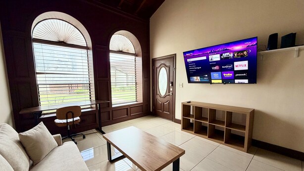 Una sala de estar con un sofá, una mesa de centro, un televisor montado en la pared y ventanas grandes con persianas.