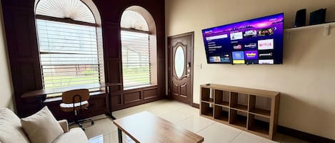 Una sala de estar con un sofá, una mesa de centro, un televisor montado en la pared y ventanas grandes con persianas.