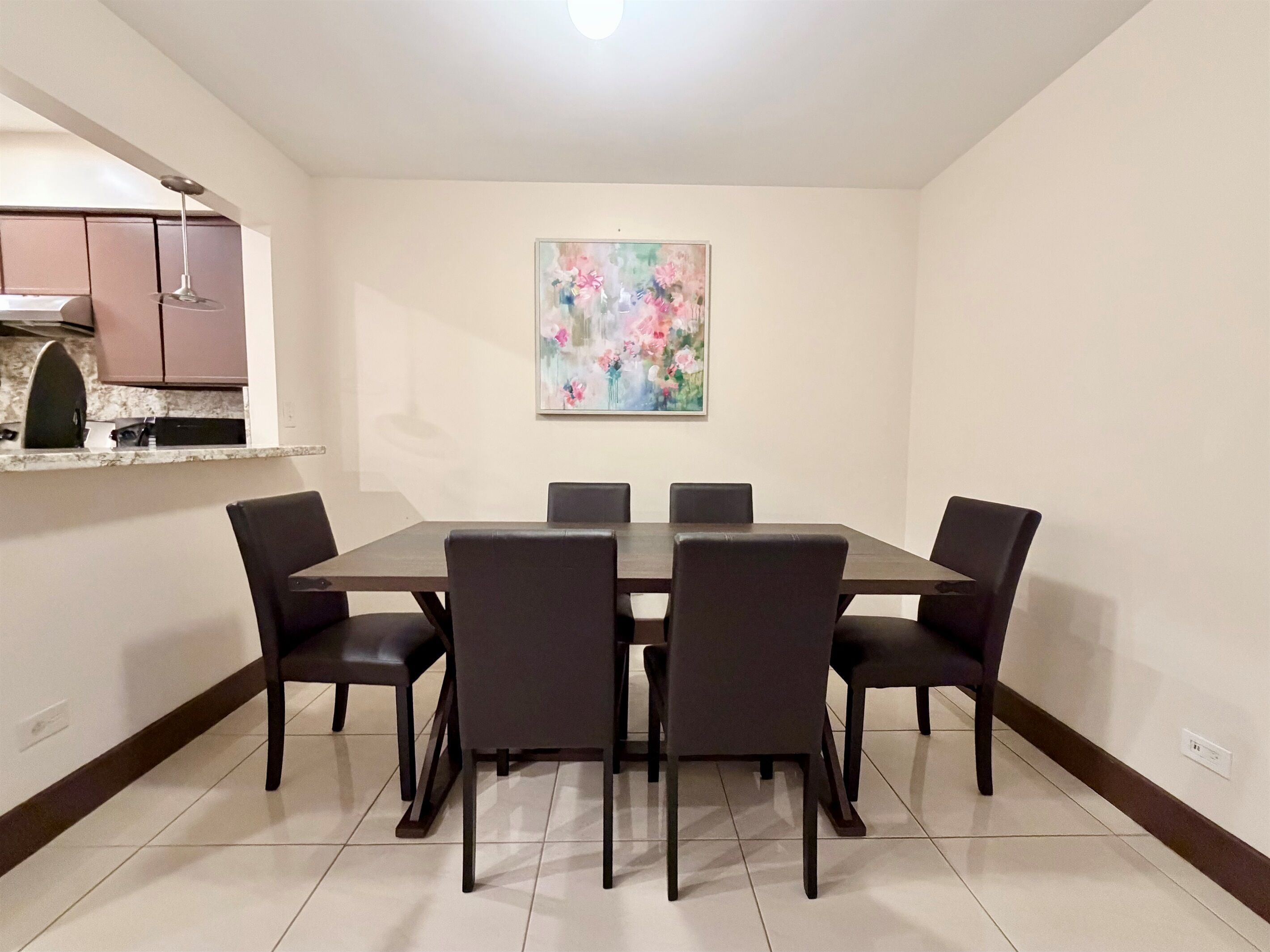 Un comedor con una mesa rectangular y seis sillas, un cuadro en la pared y una zona de cocina con armarios y una encimera.