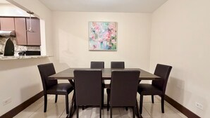 Un comedor con una mesa rectangular y seis sillas, un cuadro en la pared y una zona de cocina con armarios y una encimera.