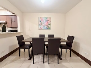 A dining area with a rectangular table and six chairs, a painting on the wall, and a kitchen area with cabinets and a countertop.