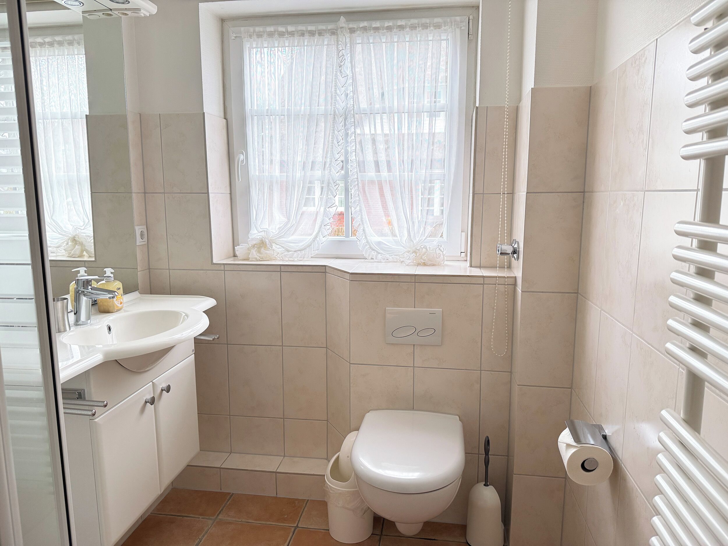 Ein Badezimmer mit Toilette, Waschbecken und einem Fenster mit transparenten Vorhängen.