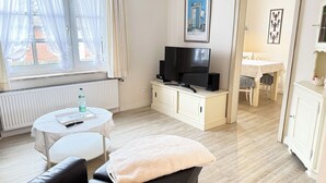 A living room with a sofa, a TV on a cabinet, a dining table with chairs, and a window with curtains. - Vacation apartment Um's Eck in Haus Inselfriede very close to the beach in Westerland (Westerland)