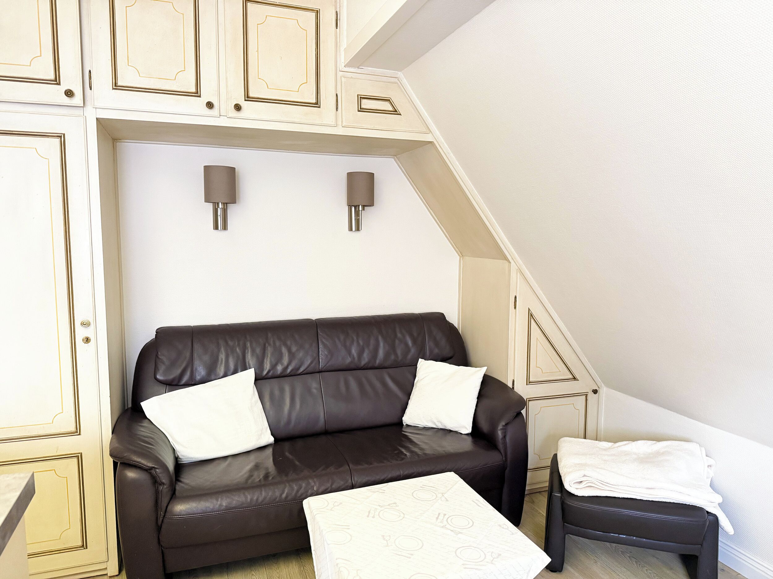 A brown leather sofa with white cushions, a small table with a white patterned tablecloth, and a chair with a white towel.