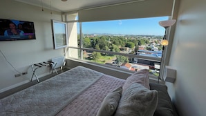 Un dormitorio con cama, ventilador de techo, televisor y un amplio ventanal con vista a la ciudad.