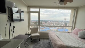 Un dormitorio con cama, escritorio, silla y vistas a un paisaje urbano a través de amplias ventanas.