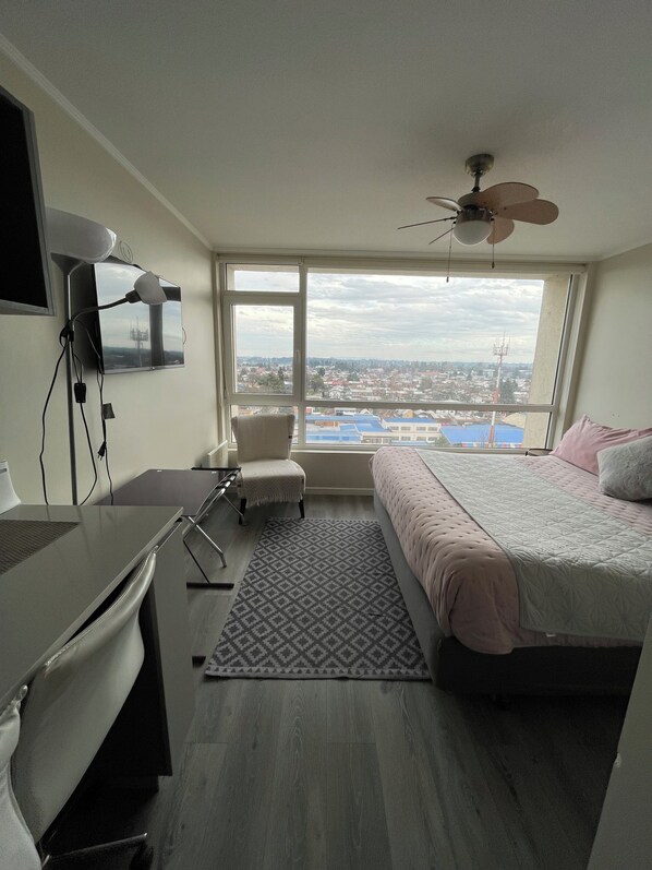 Une chambre à coucher comprenant un lit, un bureau, une chaise et offrant une vue sur le paysage urbain grâce à de grandes fenêtres.