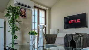 Una sala de estar con una mesa de vidrio, un sofá, un televisor mostrando 'NETFLIX' y un ventanal grande.