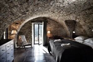 Un dormitorio con paredes de piedra, una cama, una silla, un aparador y una lámpara.