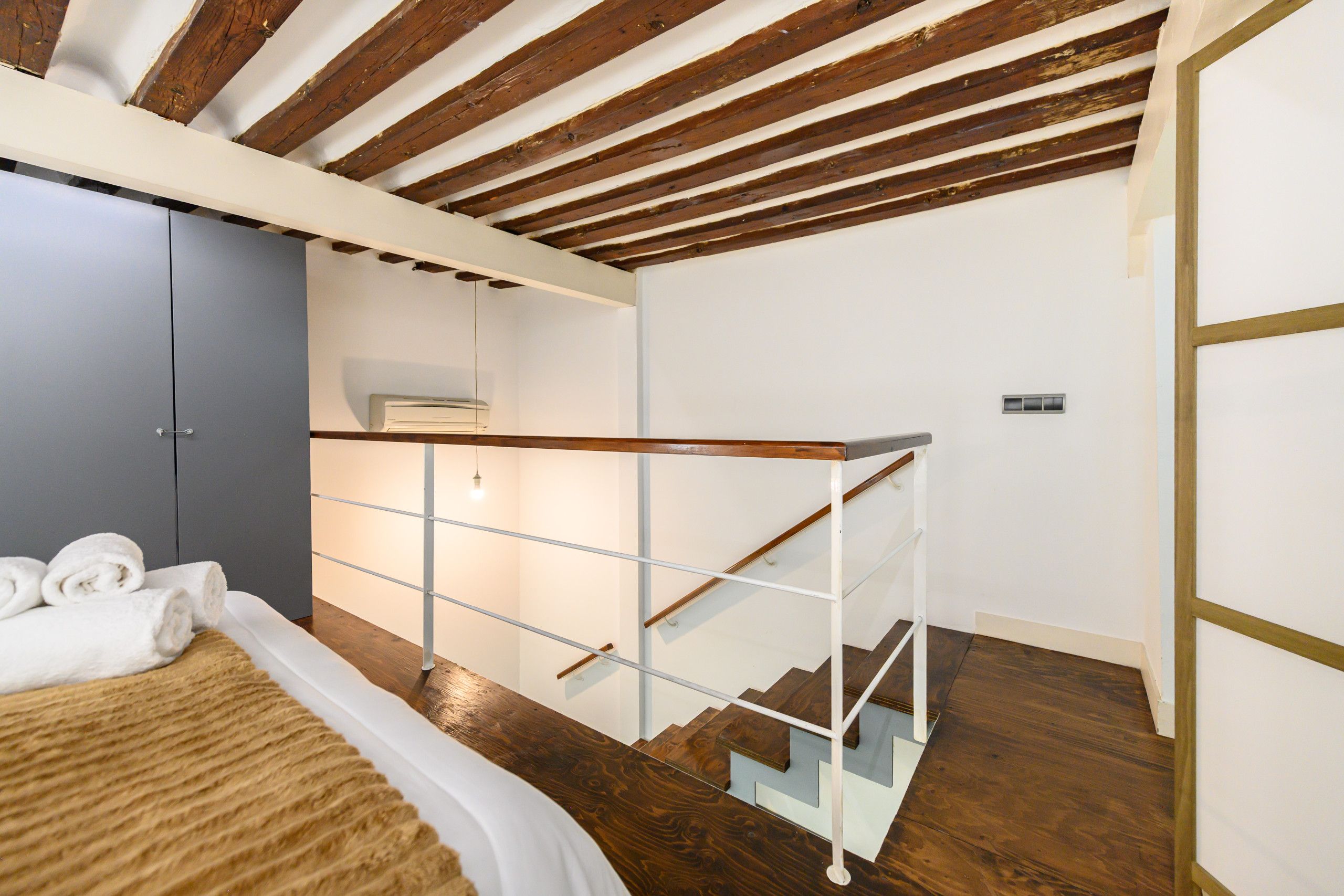 Un dormitorio con techo de madera, una cama con ropa blanca, un armario gris y una escalera con barandilla de cristal.