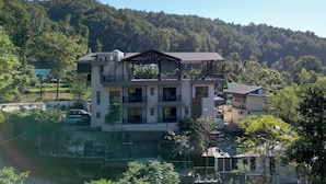 Un edificio de varios pisos con balcones, una terraza cubierta y vistas a una colina boscosa.