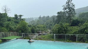 A swimming pool with a person swimming, surrounded by a fence and lush greenery. - Saraiville - Boutique Riverside Stay (Vikasnagar)