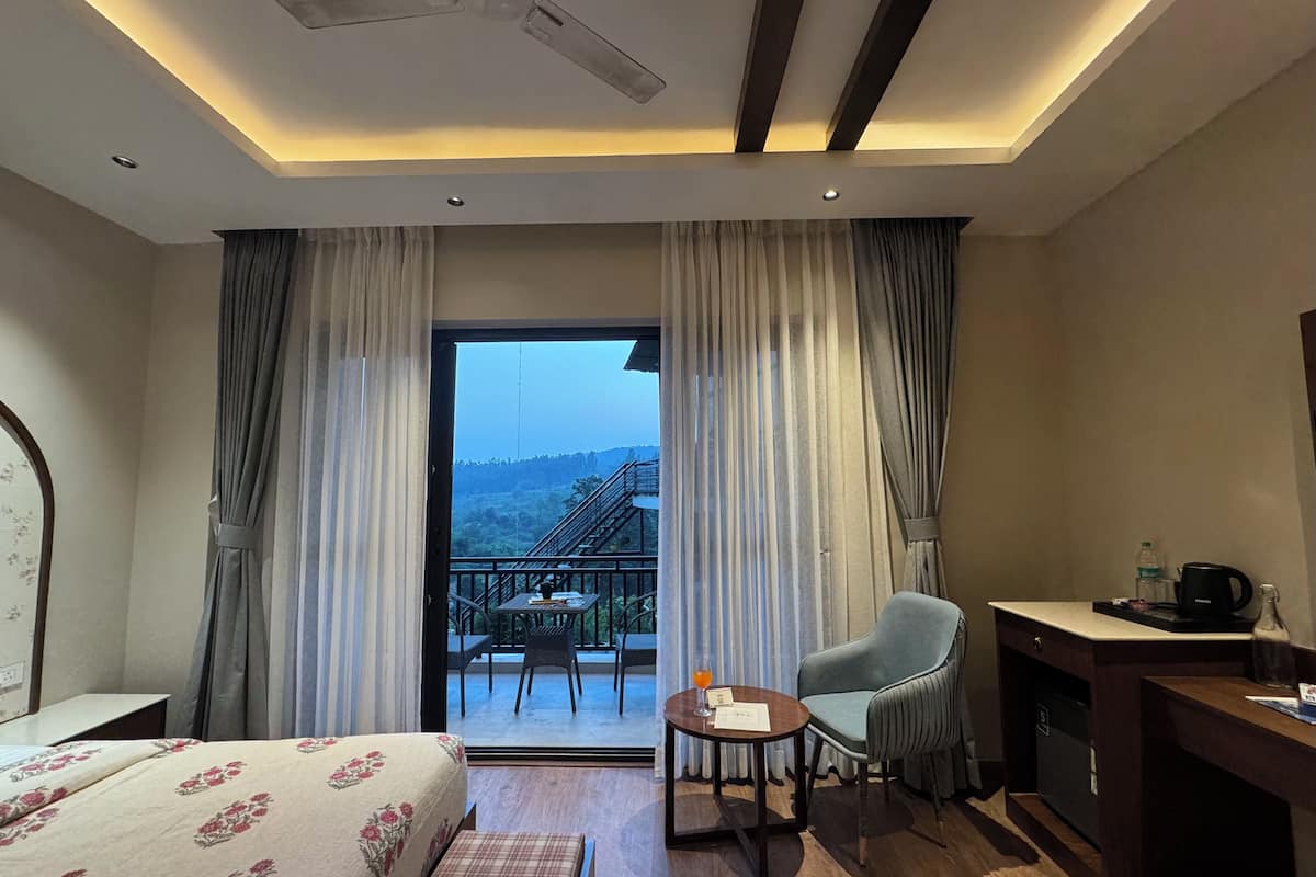 Une chambre d’hôtel comprenant un lit, un bureau, une chaise, une petite table avec un verre, et un balcon offrant une vue sur les collines.