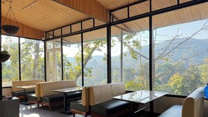 A spacious room with large windows, wooden ceiling, and a view of a mountainous landscape. - Saraiville - Boutique Riverside Stay (Vikasnagar)