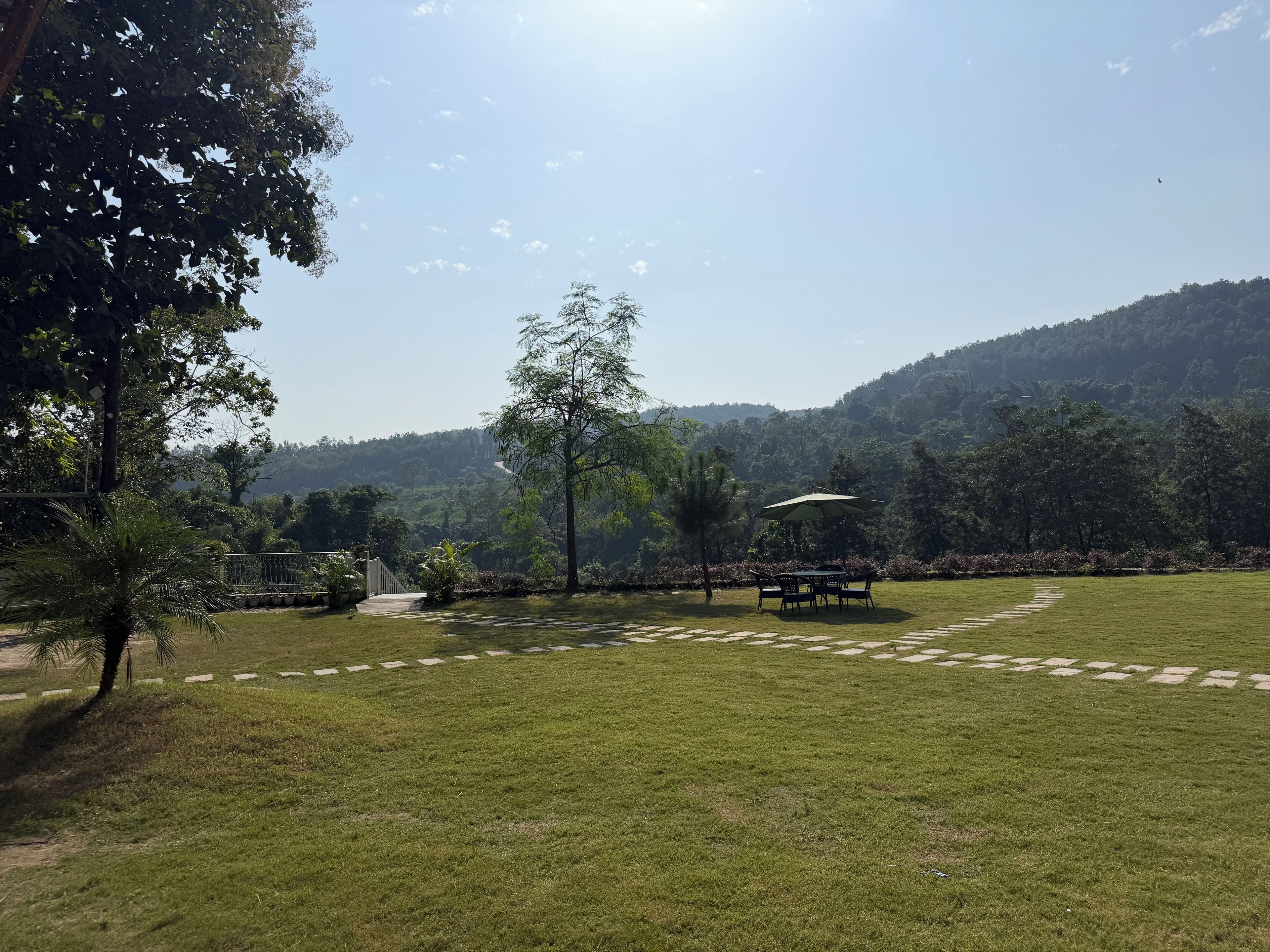A grassy outdoor area with a paved path, a table with chairs, and a large umbrella.