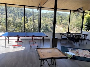 A spacious room with a ping pong table, a foosball table, and a view of a forested area through large windows. - Saraiville - Boutique Riverside Stay (Vikasnagar)