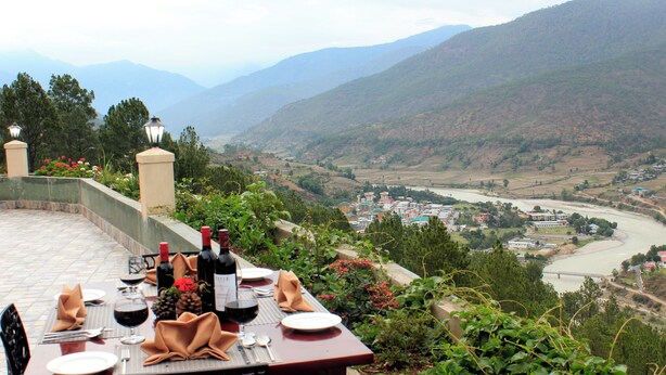 A table set for dining with wine bottles, glasses, and a view of a valley with a river and buildings. - Hotel Sonamgang (Punakha)