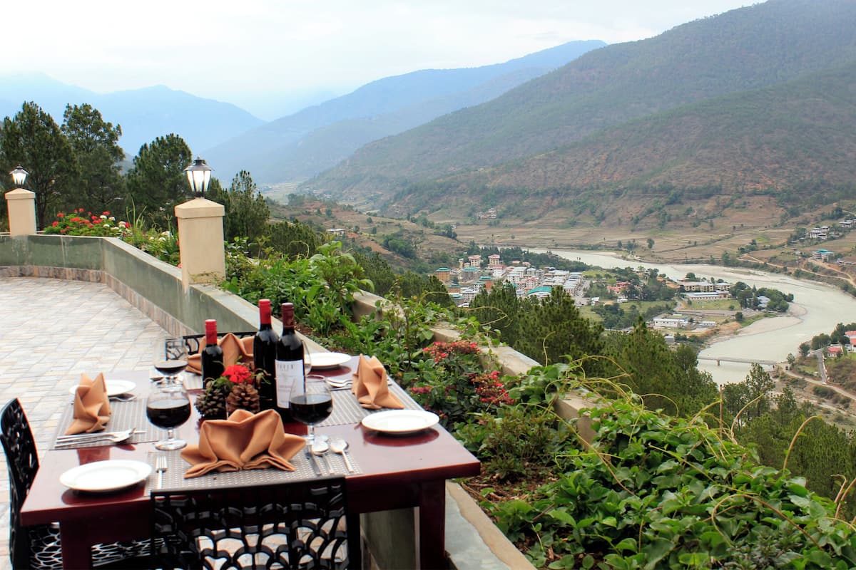 A table set for dining with wine bottles, glasses, and a view of a valley with a river and buildings.