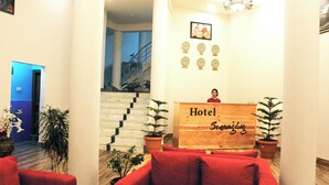 A hotel reception area with a wooden counter, red sofas, potted plants, a staircase, and a wall with clocks and a framed photo. - Hotel Sonamgang (Punakha)