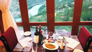 A dining table with a view of a river and hills, wine bottles, glasses, and plates of food. - Hotel Sonamgang (Punakha)