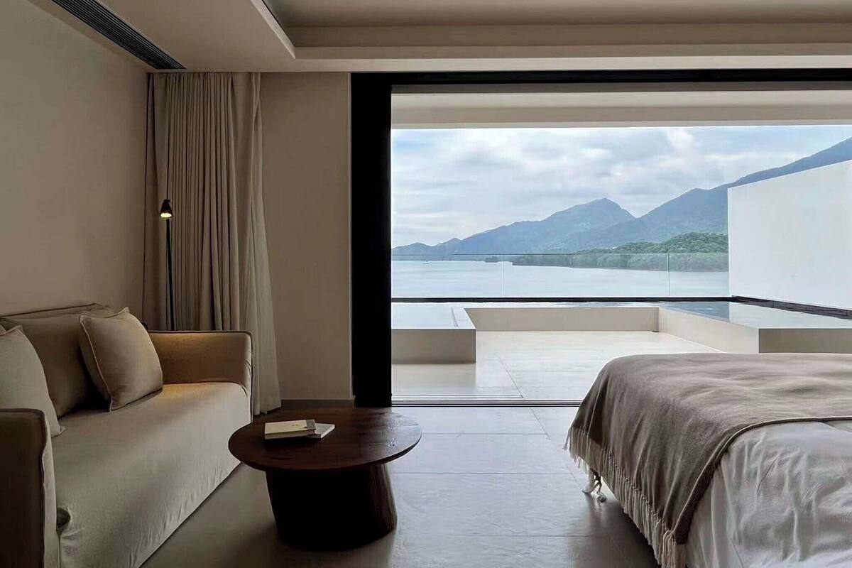 Ein modernes Hotelzimmer mit einer Couch, einem runden Couchtisch, einem Bett und einem Balkon mit Blick auf einen See und Berge.