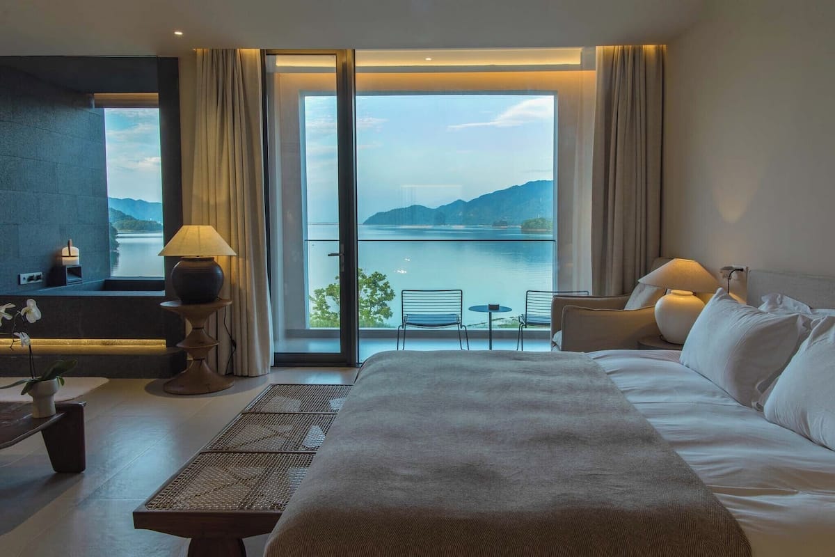 Ein Hotelzimmer mit einem großen Bett, einem Schreibtisch mit Lampe, Blick auf einen See und Berge sowie einem Balkon mit Sitzgelegenheiten.