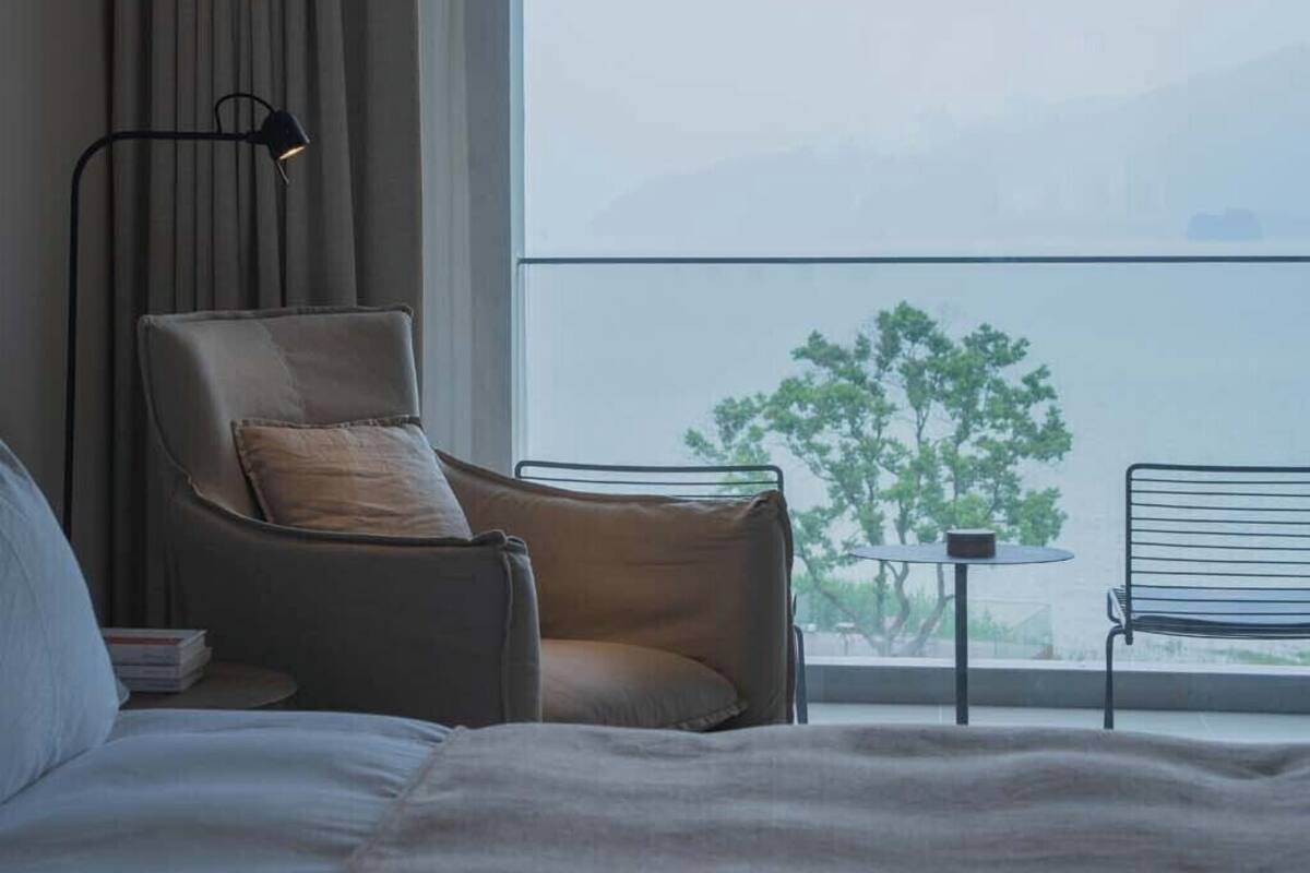 Ein Hotelzimmer mit Bett, Stuhl, kleinem Tisch und Blick auf einen Baum durch ein großes Fenster.