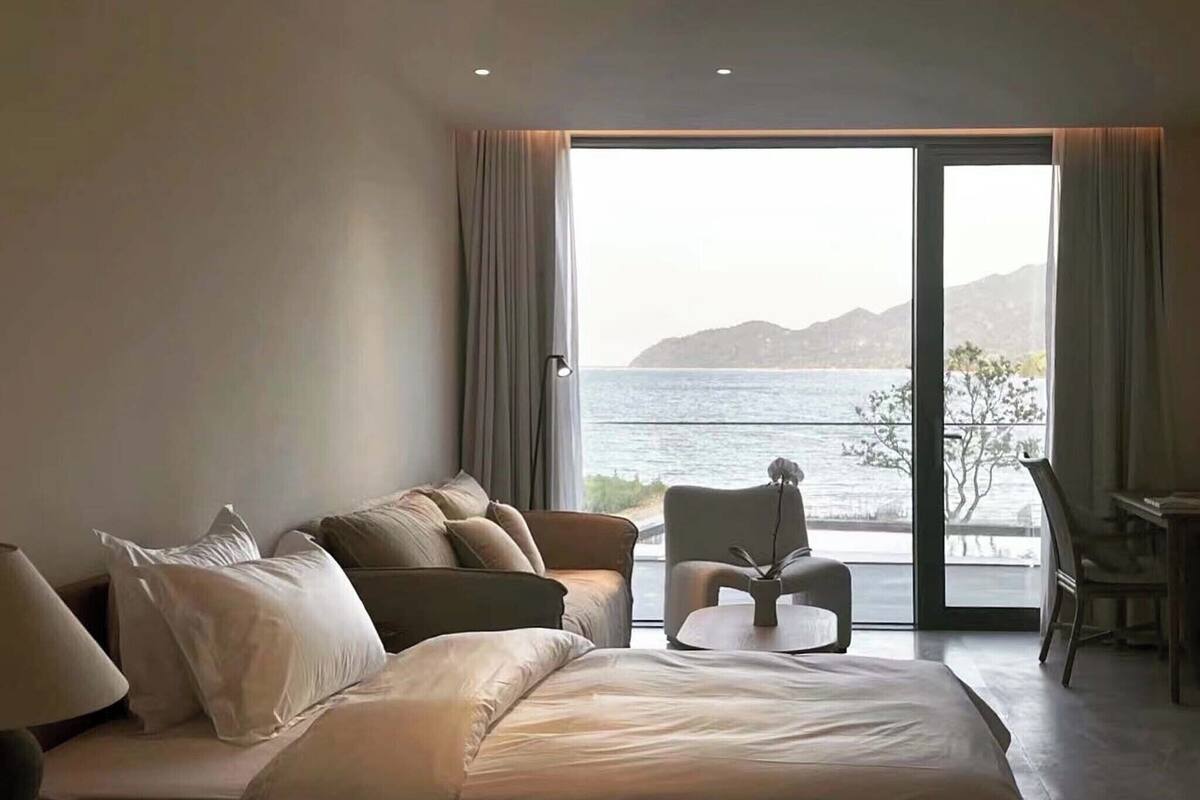 Ein Hotelzimmer mit Bett, Sofa, Sessel und Schreibtisch. Ein großes Fenster bietet Blick auf das Meer und die Berge.