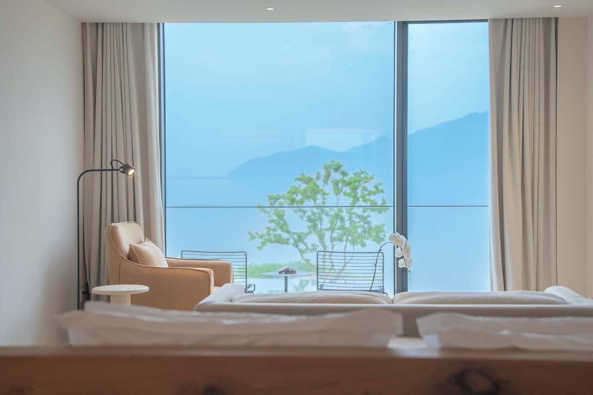 Ein Hotelzimmer mit Bett, Stuhl, kleinem Tisch und großem Fenster mit Vorhängen.