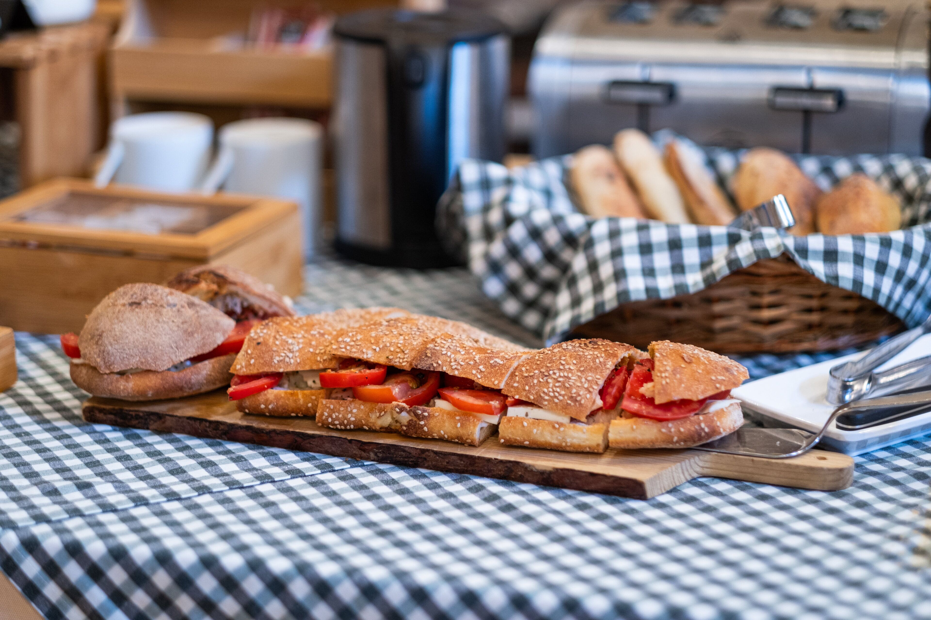A sandwich on a wooden board, a basket of bread rolls, and a coffee machine in the background.