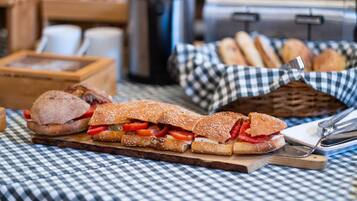 A sandwich on a wooden board, a basket of bread rolls, and a coffee machine in the background.