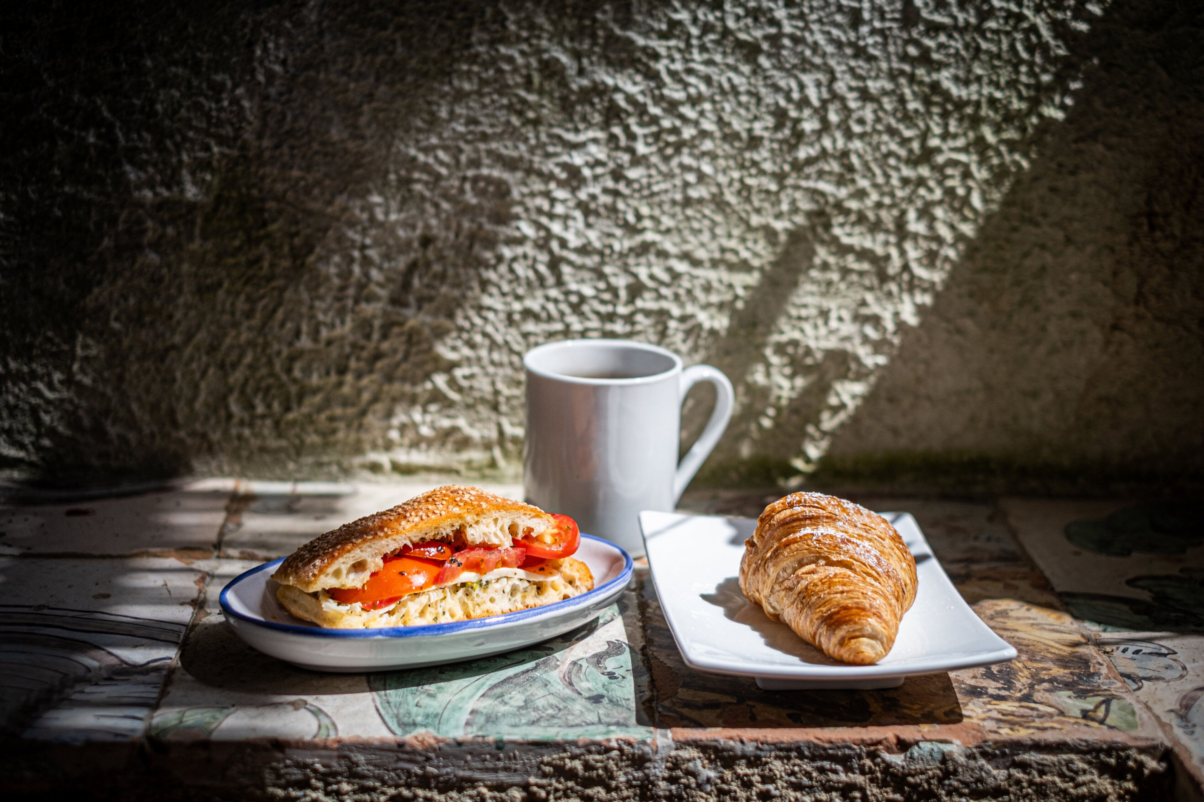 A sandwich on a plate and a croissant on a small plate, both on a table with a textured wall in the background.