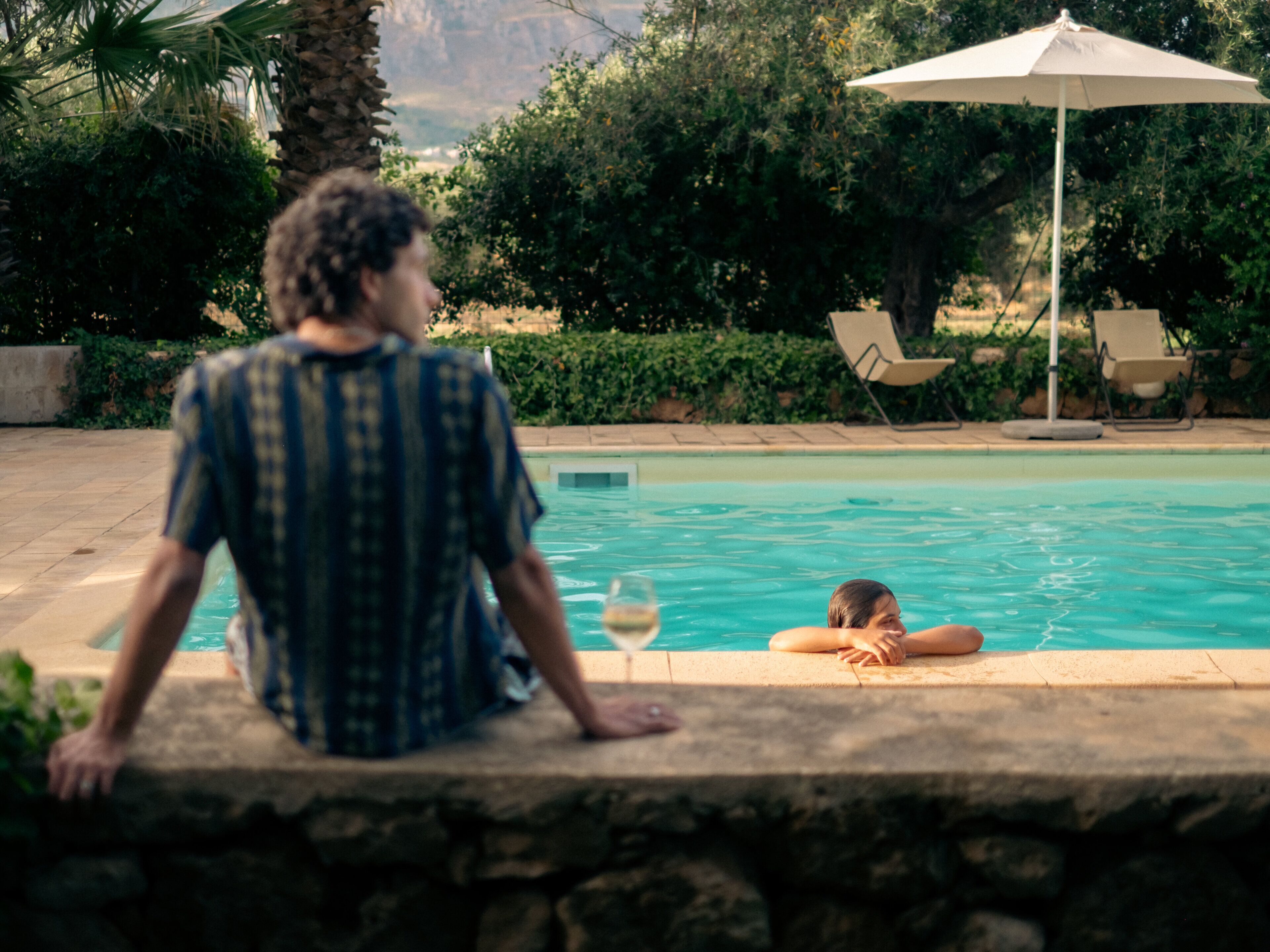 Un hombre sentado junto a una piscina, sosteniendo una bebida, con vistas a un área de piscina con sillones de descanso y una sombrilla grande.
