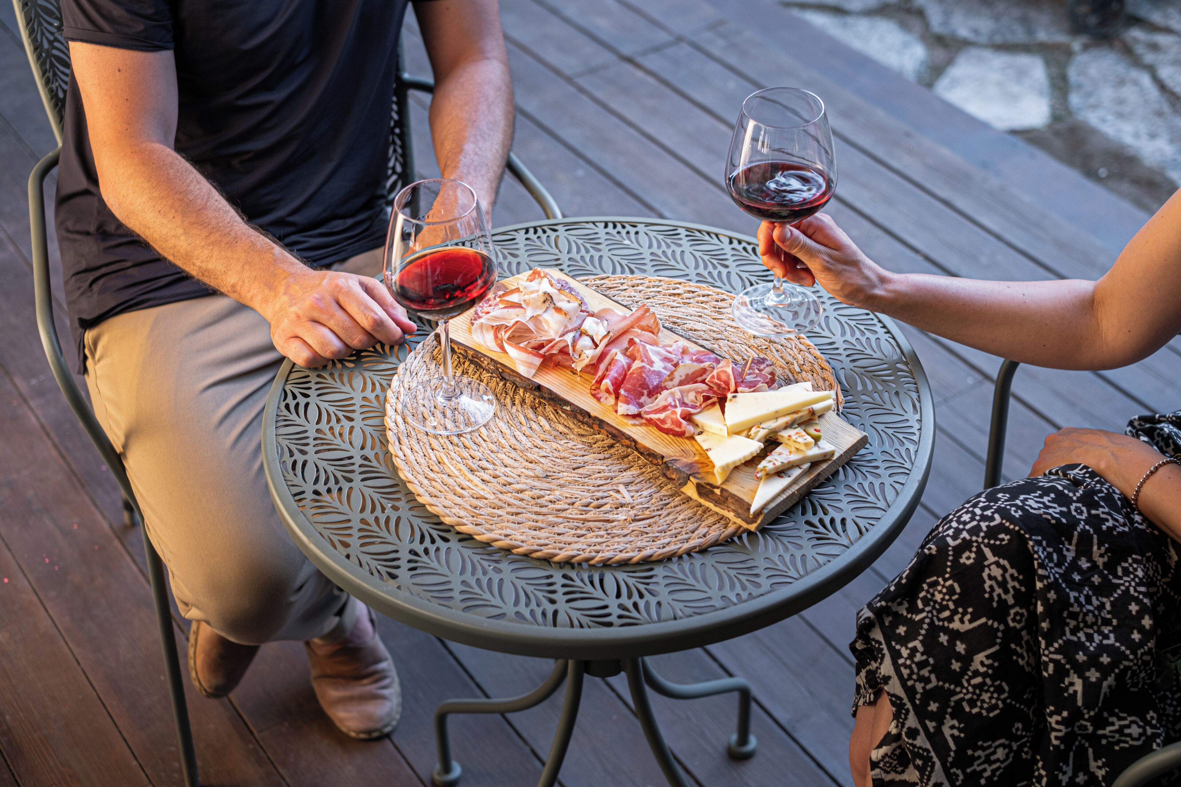 Un tavolo rotondo in metallo con un sottobicchiere intrecciato, un tagliere con salumi e formaggi, e due bicchieri da vino con vino rosso.
