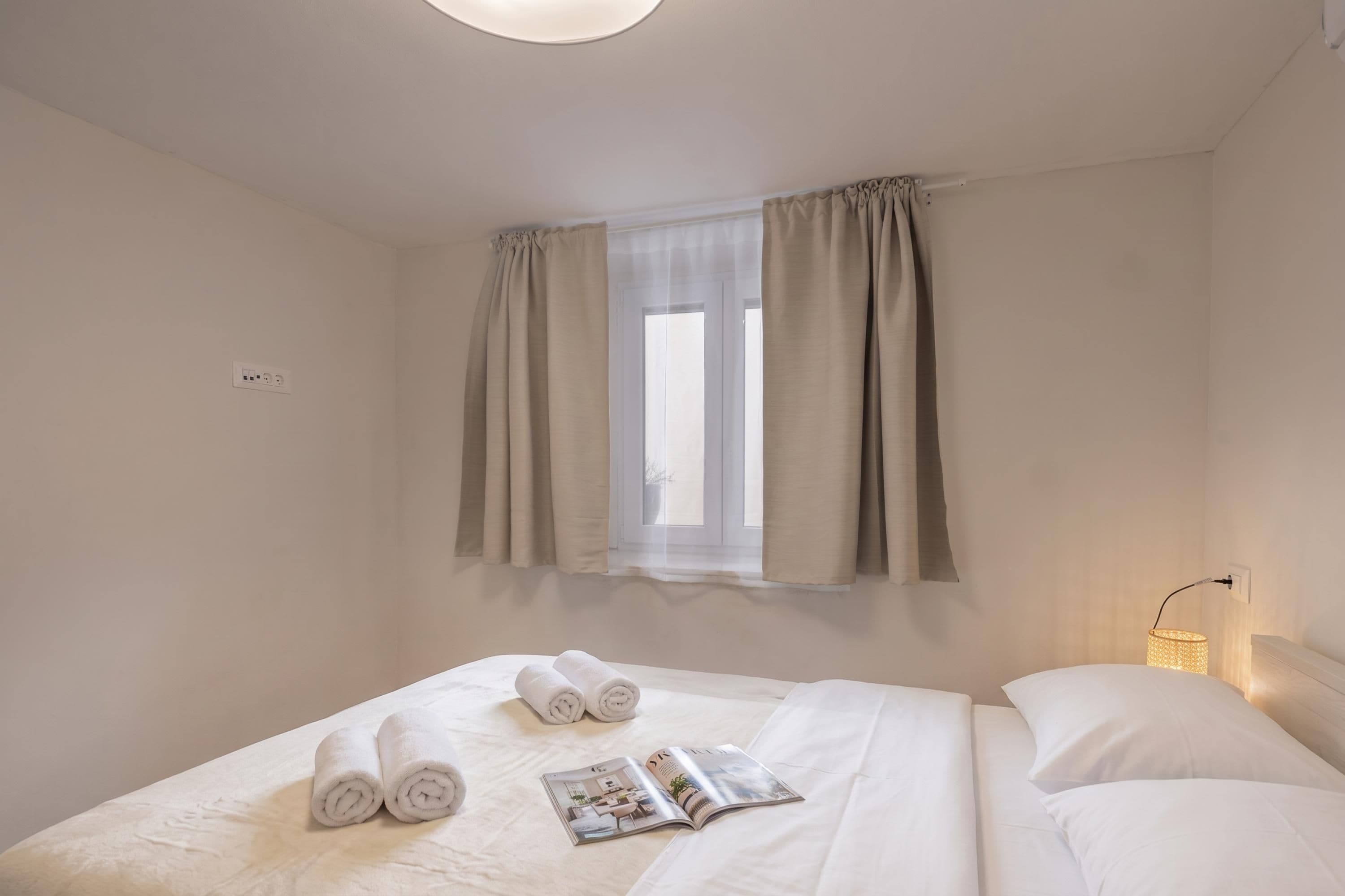 A hotel room with a bed, two rolled towels, a magazine, a window with curtains, and a bedside lamp.