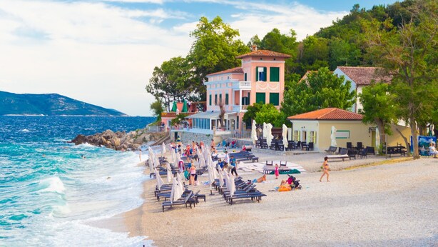 A beach with sun loungers, a building with a red roof, and a view of the sea. - Casa del Porto Seafront Apartment (Moscenicka Draga)