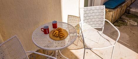 A small outdoor table with a croissant and two red cups, surrounded by white metal chairs.