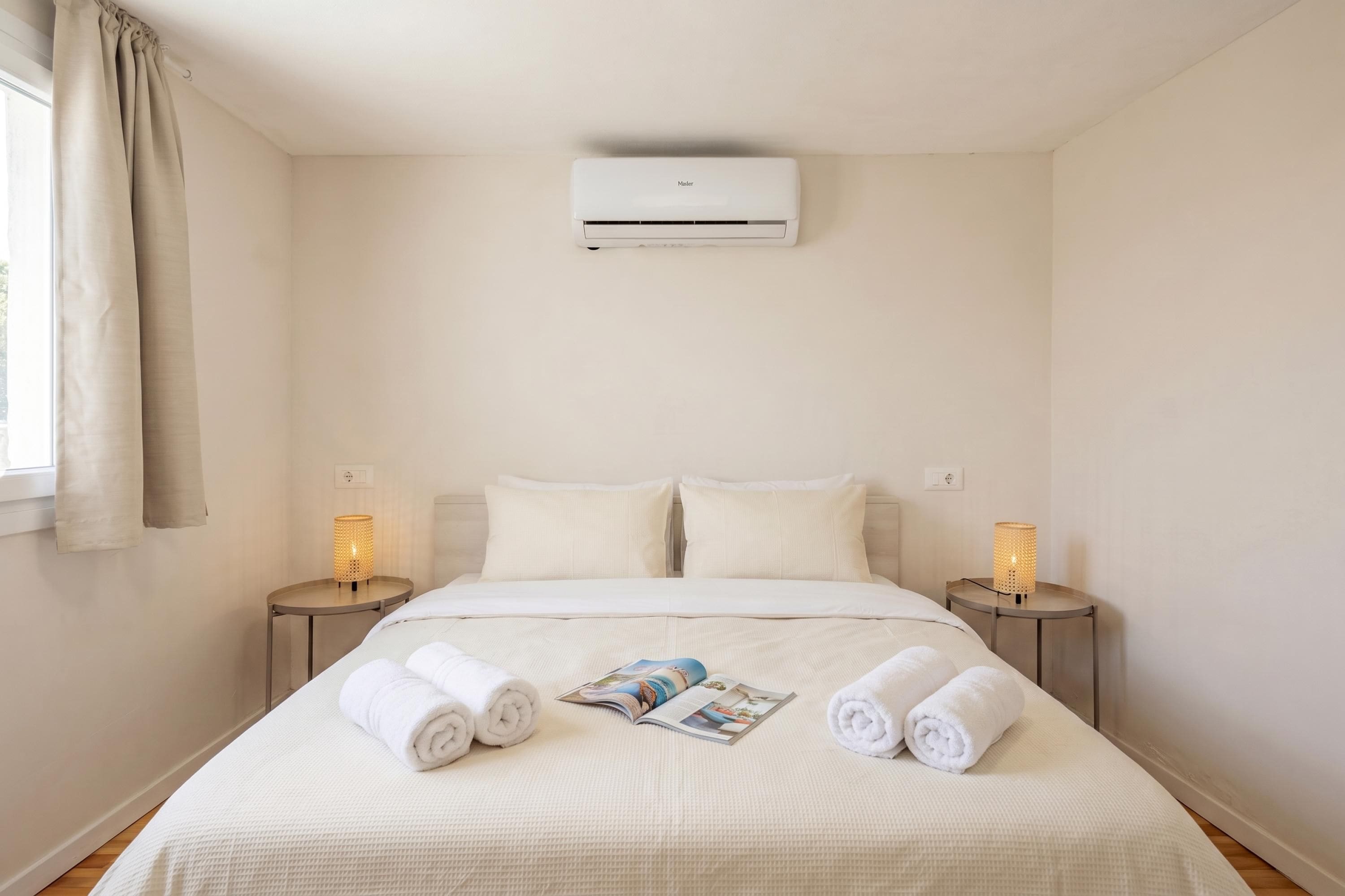 A hotel room with a bed, two bedside tables, an air conditioner, and a window with curtains.