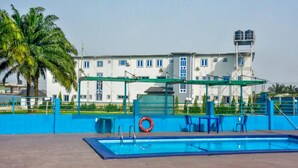 A swimming pool with a blue border, a palm tree, and a building with multiple windows and a water tower in the background. - MBL HOTEL  (Alakahia)