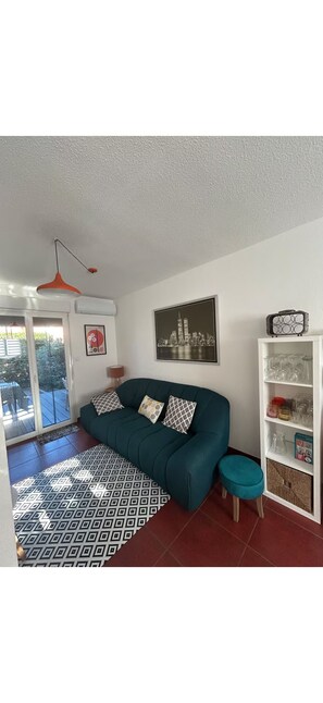 Una sala de estar con un sofá azul turquesa, un tapete estampado, una vista urbana enmarcada y una estantería blanca con cristalería.