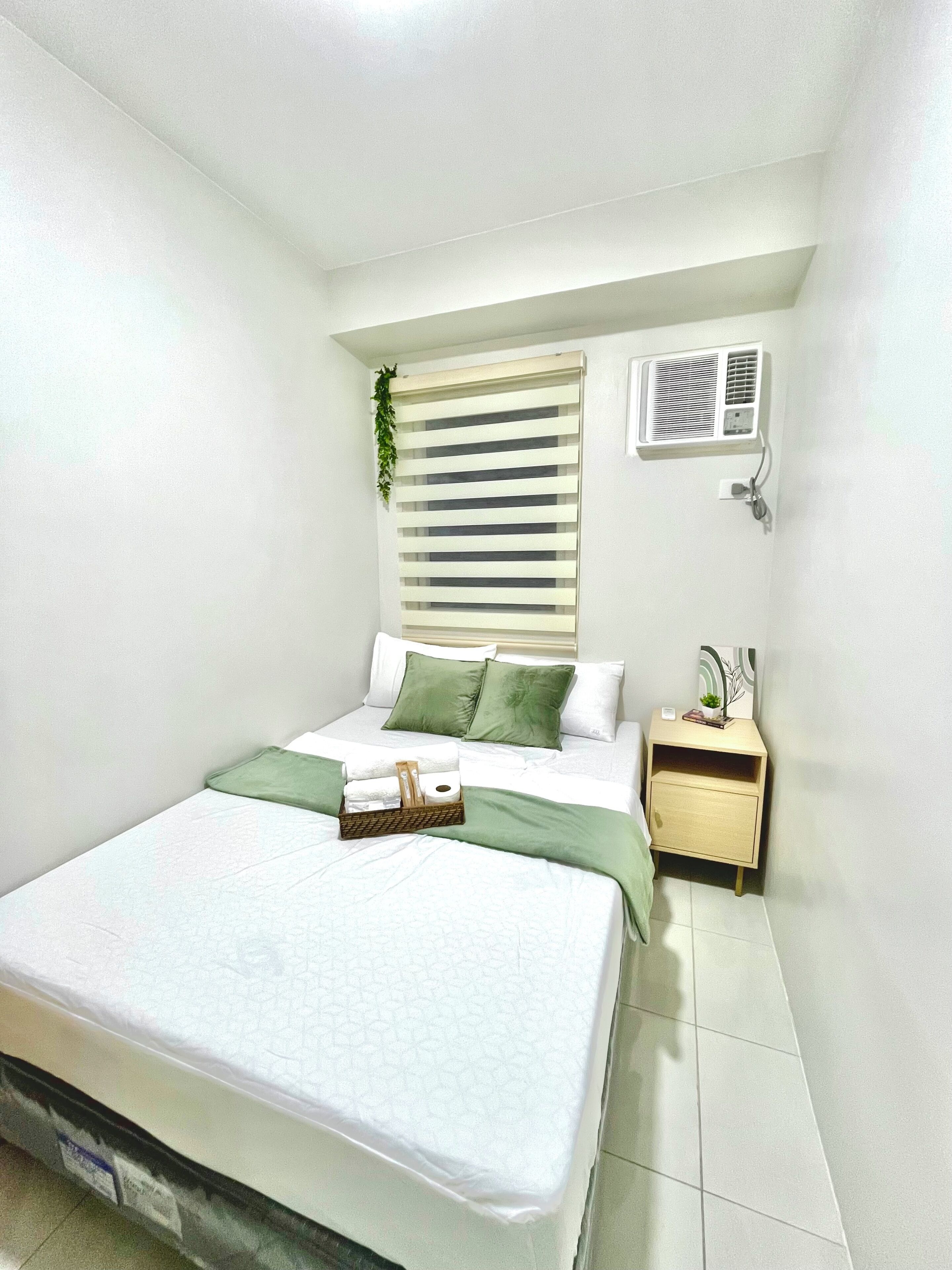 A bedroom with a bed, bedside table, nightstand, air conditioning unit, and a window with blinds.